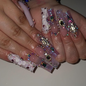 Könnte beinhalten: Lange, verzierte Kunstnägel in verschiedenen Designs. Einige Nägel weisen weiße Blumenakzente auf, während andere eine Reihe farbenfroher Strasssteine und Edelsteine zeigen. Die Nägel haben eine hellrosa Grundfarbe mit Glitzer.