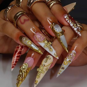May include: A set of long, pointed acrylic nails with intricate designs. The nails feature gold embellishments, including rings, crosses, and decorative accents. Colors include white, cream, red, and gold, creating a luxurious and detailed aesthetic.