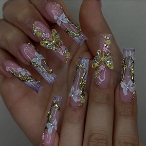 May include: Long, coffin-shaped artificial nails with a light pink base and intricate designs. Each nail features gold accents, white flowers, and sparkling rhinestones. Some nails have butterfly and floral embellishments.