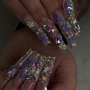 Op de afbeelding: Lange, heldere acrylnagels met ingewikkelde ontwerpen. Elke nagel heeft een glitterbasis en iriserende accenten, versierd met 3D-lavendelbloemen, zilveren vlinders en kleurrijke strass-steentjes. Een glamoureuze accessoire.