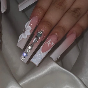 Puede incluir: Uñas acrílicas largas y cuadradas con un diseño degradado rosa claro y blanco. Algunas uñas presentan purpurina blanca, pedrería y una flor en 3D. Una uña tiene la letra "A" en blanco. Un diseño de uñas glamuroso.