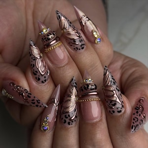 Könnte beinhalten: Eine Maniküre mit langen, spitzen Kunstnägeln. Die Nägel weisen ein Leopardenmuster, metallische Akzente und dekorative Juwelen auf. Einige Nägel haben ein gestapeltes Ringdesign, andere eine schmetterlingsförmige Verzierung.