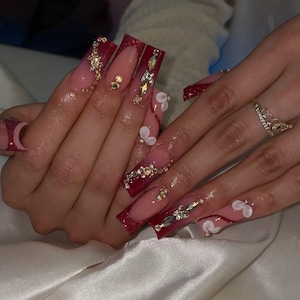 Pode incluir: Unhas artificiais quadradas com borda de glitter vermelho e gradiente rosa a vermelho. Enfeitadas com strass e pequenos detalhes florais brancos. Um anel dourado com design de coroa é visível.