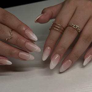 Op de afbeelding: Close-up van handen met lange, amandelvormige nagels met een French manicure design. De nagels hebben een gradiënteffect, van een nude basis naar een witte punt, omlijnd met een dunne witte lijn. Gouden ringen sieren de vingers.