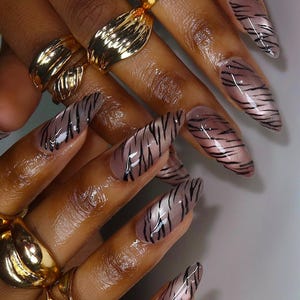 May include: Close-up of hands with long, almond-shaped nails. The nails feature a tiger stripe design in black and brown. The fingers also have gold rings.