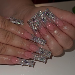 May include: Long, square-shaped artificial nails with a pink ombre effect and encrusted with iridescent rhinestones. The nails are highly embellished with various sizes and shapes of sparkling crystals, creating a glamorous and eye-catching design.