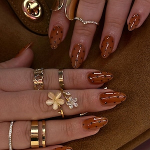 Könnte beinhalten: Nahaufnahme von Händen mit goldenen Ringen und braunem Nagellack mit schwarzen Punkten. Zu den Ringen gehören ein Blumenmuster, ein Band aus kleinen Diamanten und verschiedene goldene Bänder. Die Nägel sind oval geformt.