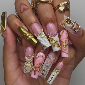May include: A hand displaying long, ornate acrylic nails. The nails feature a variety of designs, including pink and white French tips, gold embellishments, pearls, crosses, and miniature portraits. Some nails have a 444 design. Gold rings adorn the fingers.