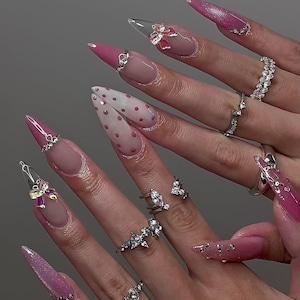Op de afbeelding: Close-up van gemanicuurde nagels met diverse ontwerpen. De nagels tonen roze, transparante en polkadot patronen, versierd met strass en strikaccenten. Verschillende zilveren ringen met edelsteenaccenten zijn ook zichtbaar.