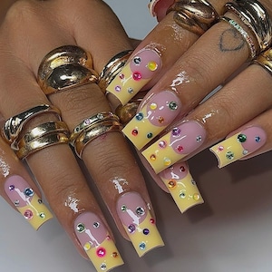 Pode incluir: Close-up de unhas acrílicas longas e quadradas. As unhas são pintadas de amarelo com uma forma de meia-lua rosa na base. Cada unha é adornada com strass coloridos. Anéis de ouro de vários designs estão nos dedos.
