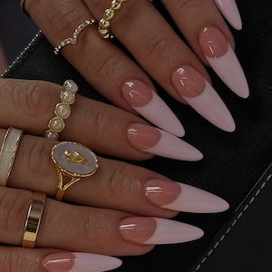 Pode incluir: Close-up de unhas longas em formato amêndoa com um design de manicure francesa. As unhas apresentam uma base rosa pálido com uma ponta nude. Vários anéis de ouro com vários designs adornam os dedos.