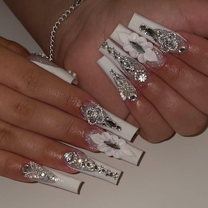 Könnte beinhalten: Lange, weiße Acrylnägel mit aufwendigen Designs. Jeder Nagel weist eine Kombination aus funkelnden Strasssteinen, silbernen Akzenten und 3D-Blütenverzierungen in Weiß auf. Die Nägel sind quadratisch geformt und haben ein glänzendes Finish.