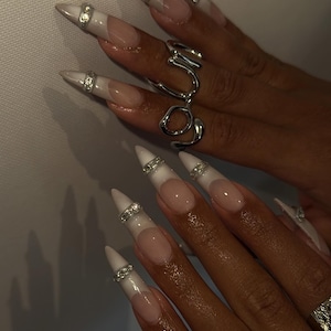 May include: Close-up of long, pointed fingernails with a white and nude design, accented with silver rhinestone bands. A silver ring with an abstract design is also visible, showcasing a modern nail art style.
