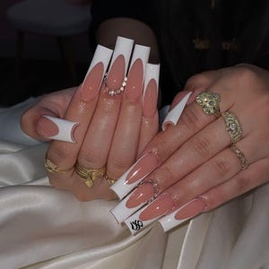 May include: Long, square-shaped acrylic nails with a French manicure design. The nails feature a white tip, a nude base, and a small rhinestone accent. The nails are adorned with multiple gold rings.
