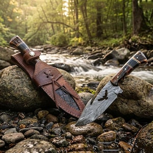 Puede incluir: Un cuchillo de caza artesanal con una hoja estampada y un mango de madera, apoyado sobre una roca junto a una funda de cuero. El cuchillo presenta un recorte de águila. La funda es marrón con textura de cocodrilo.