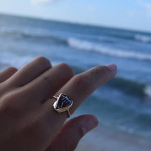Peut inclure: Une bague dorée ornée d'une pierre triangulaire brun foncé avec un motif blanc rappelant des veines, sertie dans une monture en métal lisse. La bague possède un anneau fin et poli de couleur or qui entoure le doigt avec élégance.