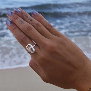 May include: A silver ring with an oval, light-coloured stone is worn on a hand with blue nail polish. The ring is the focal point, set against a blurred ocean backdrop. A gold bracelet is also visible.
