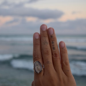 Può includere: Un anello con una conchiglia grigia di forma ovale e superficie liscia, montata su una fascia metallica. La conchiglia presenta un motivo naturale scuro simile a una spina che corre verticalmente al centro, creando un contrasto con il grigio.