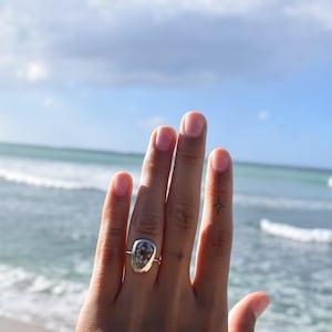 May include: A silver ring featuring an irregular, rounded bezel setting that holds a speckled, grey and white stone. The ring band is thin and polished, showcasing a smooth metallic finish that contrasts with the textured, organic appearance of the stone.