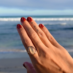 Peut inclure: Une bague ornée d'un coquillage cauri de forme ovale monté sur un anneau métallique. Le coquillage présente une surface lisse de couleur beige avec une fente verticale centrale révélant un intérieur texturé de couleur crème.