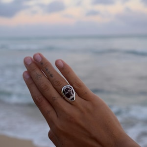 Puede incluir: Un anillo de plata con una piedra irregular en forma de lágrima que presenta un patrón jaspeado en marrón oscuro y blanco. La piedra está engastada en un bisel de plata lisa y pulida que sigue la forma orgánica y asimétrica.