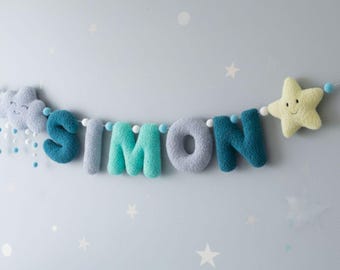 May include: A decorative garland featuring plush, textured letters spelling SIMON in shades of blue and gray, flanked by a soft gray cloud with hanging blue droplets and a pale yellow star, all strung together with small blue and white felt balls.