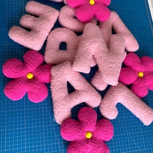 May include: A collection of plush, textured letters and flower-shaped decorations. The letters are light pink, while the flowers are vibrant magenta with small, bright yellow centers. Each piece features a soft, fuzzy, sherpa-like fabric finish.