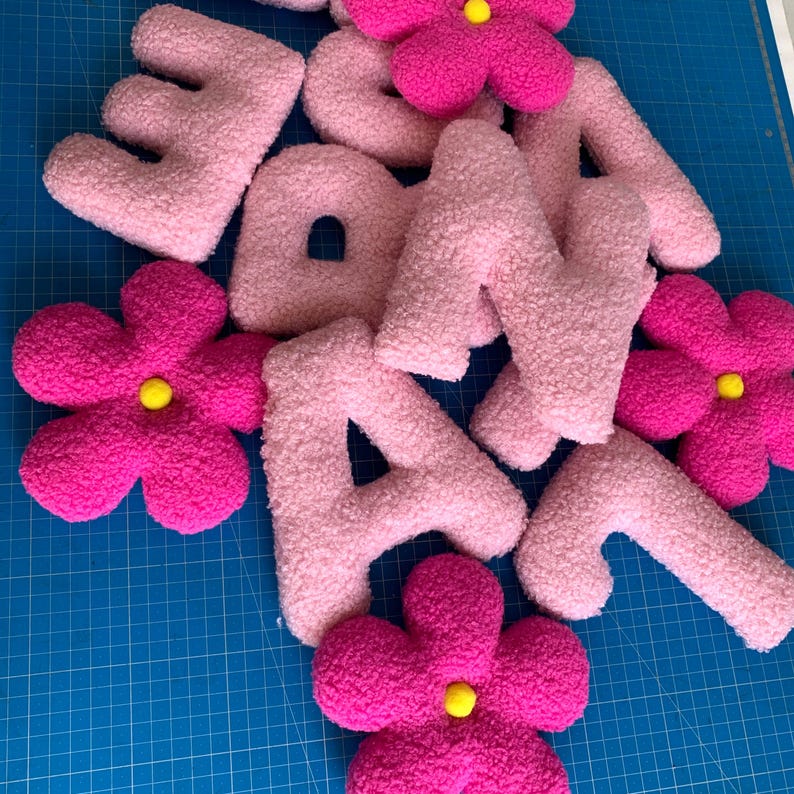 May include: A collection of plush, textured letters and flower-shaped decorations. The letters are light pink, while the flowers are vibrant magenta with small, bright yellow centers. Each piece features a soft, fuzzy, sherpa-like fabric finish.