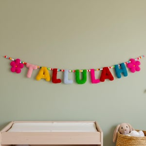 May include: A colorful felt garland spelling the name TALLULAH in individual plush letters. The letters are in shades of pink, yellow, red, light blue, green, and dark blue, interspersed with small multicolored felt balls and two large pink flowers.