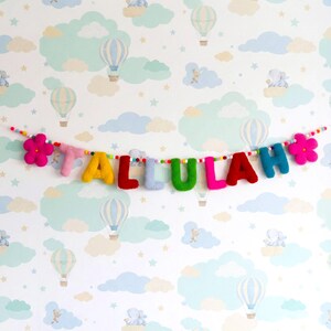 May include: A felted wool garland spelling the name TALLULAH in multicolored letters, featuring a pink flower at each end. The letters are strung on a thin cord interspersed with small, multicolored felt balls throughout the length.