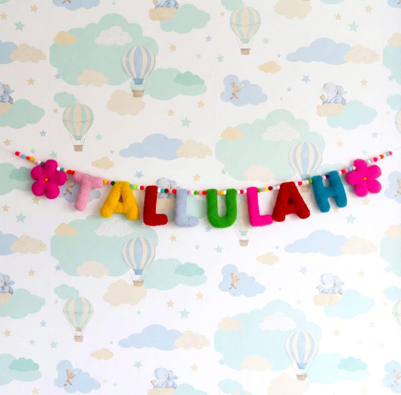 May include: A felted wool garland spelling the name TALLULAH in multicolored letters, featuring a pink flower at each end. The letters are strung on a thin cord interspersed with small, multicolored felt balls throughout the length.