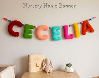 May include: A colorful nursery name banner spelling "CECILIA" in plush, textured letters. The letters are red, green, peach, turquoise, pink, yellow, and orange. The banner is strung with small, colorful pom-poms. The text "Nursery Name Banner" is above the name.