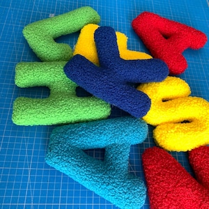 May include: A collection of plush, textured letters in vibrant colors including red, yellow, green, blue, and teal. The letters have a fuzzy, sherpa-like surface and are arranged in a scattered, overlapping pile on a blue grid surface.