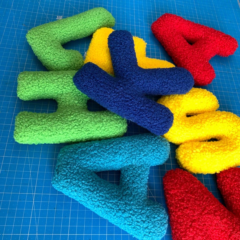 May include: A collection of plush, textured letters in vibrant colors including red, yellow, green, blue, and teal. The letters have a fuzzy, sherpa-like surface and are arranged in a scattered, overlapping pile on a blue grid surface.