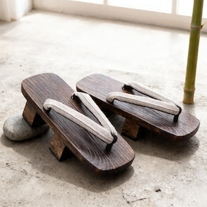 Puede incluir: Un par de sandalias geta japonesas tradicionales. Las sandalias de madera son de color marrón oscuro con una veta texturizada, con una plataforma elevada y correas de tela blanca. Las sandalias se exhiben sobre una superficie gris claro.