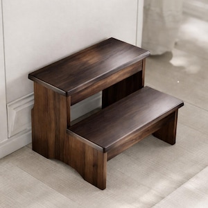 Puede incluir: Un taburete de madera de dos escalones con un acabado marrón oscuro. El taburete tiene forma rectangular con una parte superior plana y un escalón inferior, diseñado para ayudar a alcanzar lugares más altos. Se coloca contra una pared blanca.