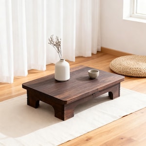 May include: A dark brown, low wooden table with a rectangular top and short legs. A small white vase with dried flowers and a small gray bowl sit on the table. The table is on a white runner.