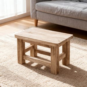 May include: A small, rectangular wooden stool with a natural finish. The stool has four sturdy legs and a simple, minimalist design. The dimensions are approximately 12 inches long, 8 inches wide, and 10 inches tall. It is placed on a beige rug.