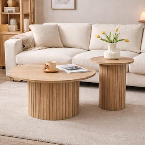 Puede incluir: Dos mesas de centro de madera con acabado marrón claro. La mesa más grande es redonda con una base cilíndrica hecha de listones de madera verticales. La mesa más pequeña también es redonda con una base similar. Un sofá blanco y una alfombra están en el fondo.