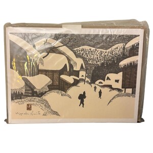 May include: Black and white print of a snowy village scene. The artwork depicts buildings covered in snow, with figures walking through the snow. The print is in a cardboard frame and wrapped in plastic.