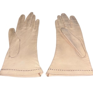 May include: A pair of light beige gloves. The gloves are made of a soft material and have a classic design. The gloves have a stitched detail along the wrist. The gloves are a neutral color.