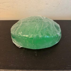 Puede incluir: Una pastilla de jabón redonda y verde envuelta en plástico transparente. El jabón tiene un aspecto liso y translúcido y está sobre una superficie oscura. El jabón es de forma ovalada.