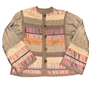 Può includere: Giacca vintage multicolore con collo tondo e maniche lunghe. La giacca presenta strisce orizzontali beige, rosa e marrone, con cuciture decorative e bottoni in legno. Le maniche hanno motivi circolari ricamati.