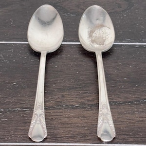 以下が含まれることがあります： 楕円形のボウルと装飾的なハンドルディテールを備えた2つのシルバースプーン。スプーンは、暗い木製の表面に並べて配置されています。スプーンはヴィンテージの美学を持っており、アンティークまたはヴィンテージの銀器である可能性があります。