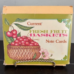 Può includere: Una scatola rettangolare di biglietti d'auguri del marchio Current con un'illustrazione di un cesto intrecciato pieno di ciliegie rosse e foglie verdi. Il testo sulla scatola riporta Current, FRESH FRUIT BASKETS e Note Cards, con fiori rosa in basso.