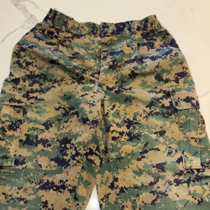 Könnte beinhalten: Tarnfarbene Cargohose in Grün-, Braun- und Schwarztönen. Die Hose hat mehrere Taschen und einen Knopfverschluss. Der Stoff hat ein pixeliges Muster, typisch für Kleidung im Militärstil.