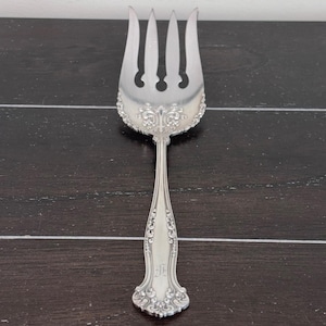 May include: A silver serving fork with four tines and a decorative floral design around the tines. The handle is slender with engraved floral patterns and a monogram. The fork is made of a shiny, reflective metal.