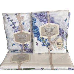 May include: A Wamsutta Supercale Plus bedding set featuring a white background with a purple and blue floral pattern. The set includes a double fitted sheet, a double flat sheet, and two standard pillowcases with Christian Dior branding.
