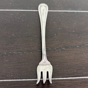Puede incluir: Un tenedor pequeño de plata con tres púas. El mango es delgado y se estrecha hacia una parte superior decorativa con la palabra "Sterling" grabada. El tenedor es probablemente para servir o una porción pequeña.