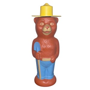 以下が含まれることがあります： ヴィンテージのスモーキーベアの置物。黄色の帽子と青いズボンを身に着け、青いシャベルを持っている茶色のクマが特徴です。「SMOKEY」の文字がベルトにプリントされています。森林火災予防のマスコットの古典的な表現です。